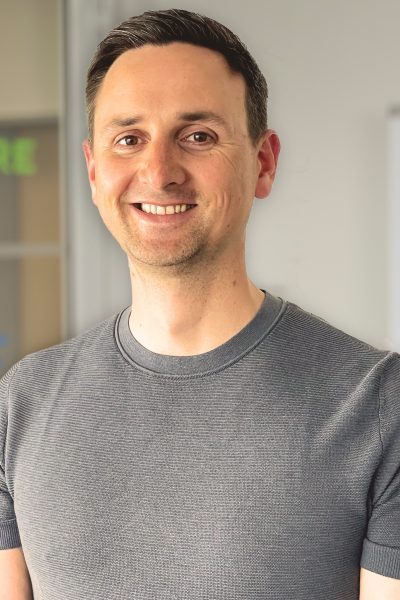 Ein freundliches Porträtfoto eines lächelnden Mannes mit kurzen, dunklen Haaren und einem dezenten Bart. Er trägt ein helles T-Shirt und blickt direkt in die Kamera. Der Mann ist Daniel Sprenger, Prokurist des Intensivpflegedienstes Kompass GmbH.