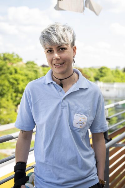 Ein Porträt einer lächelnden Person mit kurzen, hellen Haaren und Piercings im Ohr. Sie trägt ein hellblaues Poloshirt und blickt mit einem offenen Ausdruck in die Kamera. Die Person ist Dzenita Dzindo, Pflegedienstleitung des Intensivpflegedienstes Kompass GmbH