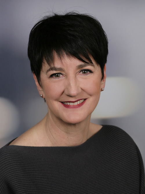 Ein Porträt von Julia Rothe mit kurzen, dunklen Haaren, die eine helle Bluse trägt. Sie sitzt entspannt in einem Sessel und blickt den Betrachter freundlich an.  Julia Rothe, Prokuristin des Intensivpflegedienstes Kompass GmbH.