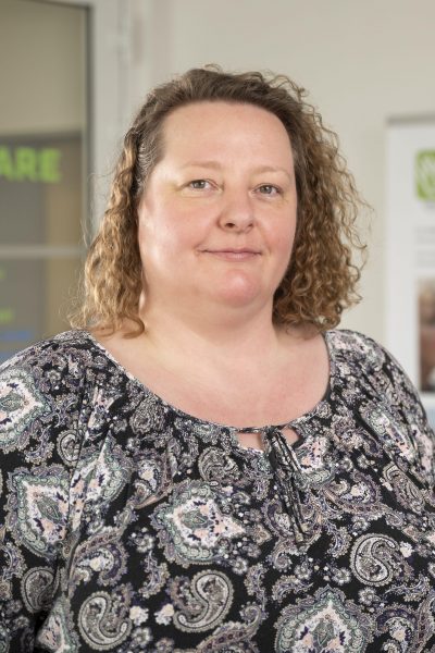 Ein Porträt einer freundlich blickenden Frau mit lockigen, hellbraunen Haaren, die über die Schultern fallen. Sie trägt ein dunkles Oberteil und blickt den Betrachter direkt an. Die Frau ist Peggy Ehrensperger, Leitung Qualitätsmanagement des Intensivpflegedienstes Kompass GmbH.